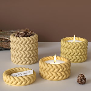 May include: Four yellow candles with a braided design. Two candles are lit, and one has a small card that says "Welcome!". One candle holder is filled with small pinecones. A decorative and cozy home decor item.