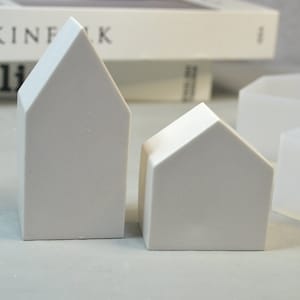 May include: Two white ceramic house-shaped candle holders, one larger than the other, on a white surface.