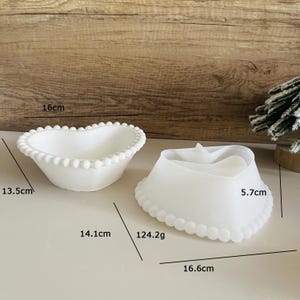 Może przedstawiać: Dwa białe ceramiczne talerze w kształcie serca z perłowymi krawędziami. Większy talerz ma 16,6 cm szerokości i 5,7 cm wysokości. Mniejszy talerz ma 14,1 cm szerokości i 13,5 cm długości.