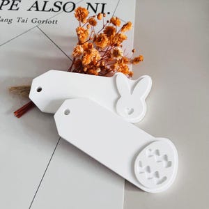 May include: Two white rectangular gift tags with a hole at the top. One tag has a bunny rabbit with a heart cutout. The other tag has a heart-shaped egg with a pattern of hearts.