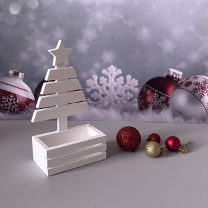 Può includere: Una fioriera a forma di albero di Natale in legno bianco con una stella in cima. Ornamenti e decorazioni glitterati rossi e dorati sono disposti nelle vicinanze. Lo sfondo presenta decorazioni a fiocco di neve e altri ornamenti natalizi.