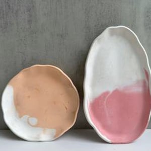 May include: Two ceramic serving dishes with an abstract design. The larger dish is oval shaped and has a white top with a pink bottom. The smaller dish is round and has a peach top with a white bottom.