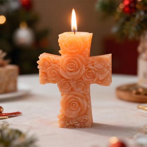 May include: A lit, peach-coloured candle in the shape of a cross, decorated with detailed rose and floral carvings. The candle is set on a white surface, with a blurred background featuring Christmas decorations.