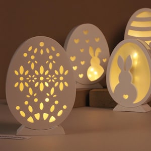 Może przedstawiać: Kolekcja białych, dekoracyjnych świateł w kształcie jajek. Każde jajko ma wycięte wzory, w tym wzory kwiatowe, serca i sylwetki królików. Światła emitują ciepłą, zachęcającą poświatę, idealną na Wielkanoc lub wiosenną dekorację.