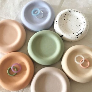 May include: Seven ceramic dishes in various colours, including light blue, white with black speckles, light green, light brown, and light pink. Each dish has a slightly raised rim and is designed to hold small items, such as rings or jewellery.