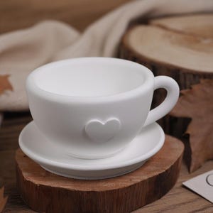 May include: White ceramic teacup and saucer with a heart design. The cup and saucer are sitting on a wooden surface.
