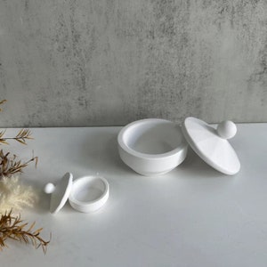 Può includere: Due contenitori in ceramica bianca con coperchi. Il contenitore più grande è aperto e quello più piccolo è chiuso. Entrambi i contenitori hanno una piccola manopola sulla parte superiore del coperchio.