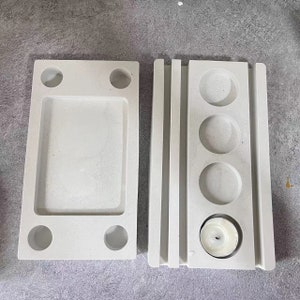 May include: Two white concrete molds for making candles. The mold on the left has a rectangular shape with a smaller rectangle inside and four round holes. The mold on the right has a vertical design with four round holes and a larger round hole at the bottom.