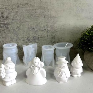 Könnte beinhalten: Eine Sammlung weißer Harzformen und -figuren. Die Formen haben die Form von Geschenktüten, während die Figuren einen Weihnachtsmann, einen Engel, einen Schneemann und einen Zwerg umfassen. Eine kleine Topfpflanze befindet sich im Hintergrund.