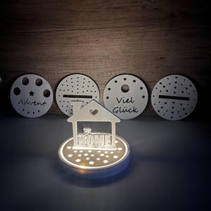 Könnte beinhalten: Weißes, hölzernes Nachtlicht in Form eines Hauses mit dem Wort "HOME" in der Mitte und einem Herz darüber. Das Licht steht auf einem runden Sockel mit Sternenmuster und ist umgeben von vier kleineren weißen Holzkreisen mit verschiedenen Designs. Die Kreise sagen "Advent", "Viel Glück" und haben ein Sternen- und Herzmuster.
