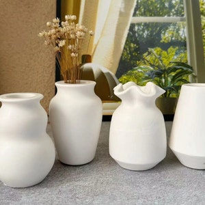May include: Four white ceramic vases of different shapes and sizes. The vases are arranged on a light grey surface.
