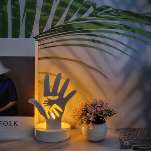 May include: White wooden night light shaped like two hands holding each other. The light is on and casting a warm glow. The night light is sitting on a table next to a book and a potted plant.