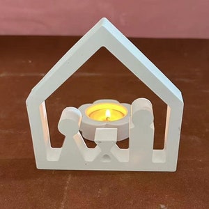 May include: White wooden candle holder shaped like a house with a cutout of a family inside. A small tea light candle is burning inside the holder.