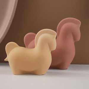 May include: Two ceramic horse figurines, one in a light brown colour and the other in a dusty rose colour. The figurines are stylised and have a simple, modern design.