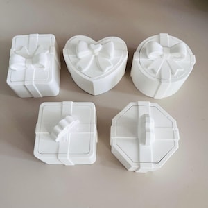 May include: Five white ceramic trinket boxes with different designs. The boxes are shaped like a heart, a square, a circle, and two different styles of a square. Each box has a raised design on the lid.