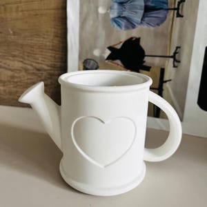 May include: White ceramic watering can with a heart design. The watering can has a spout and a handle.