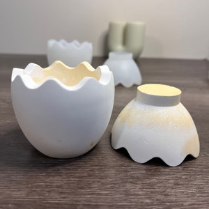 Può includere: Un set di vasi in ceramica bianca a forma di uovo con un bordo ondulato. I vasi sono separati, mostrando l'interno di un vaso.