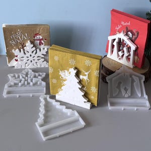 Puede incluir: Servilleteros y servilletas con temática navideña. Los soportes son blancos y tienen forma de copo de nieve, árbol de Navidad y escena de la Natividad. Las servilletas presentan diseños festivos y las palabras "Feliz Natal".