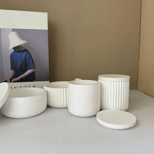 Puede incluir: Cuatro recipientes de cerámica blanca con tapas. Dos de los recipientes tienen un diseño acanalado, y los otros dos son lisos. Los recipientes están dispuestos sobre una superficie de color marrón claro.