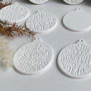 Puede incluir: Adornos de cerámica blanca con varias frases de temática navideña, incluyendo "Deck the Halls", "Home Sweet Home", "Have Yourself a Merry Little Christmas" y "It's the Most Wonderful Time of the Year".