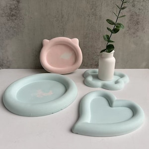 May include: Four decorative trays made of light blue and pink concrete. The trays are shaped like a heart, a circle, a cloud, and a cat's face.