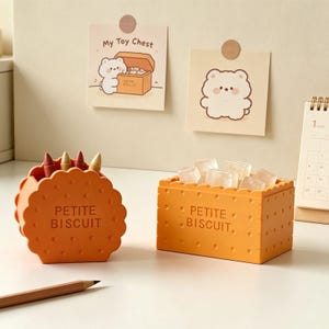 Puede incluir: Dos recipientes naranjas con forma de galleta, uno con crayones y el otro lleno de cubitos de hielo, sobre una superficie blanca. El texto "PETITE BISCUIT" está impreso en ambos recipientes. Dos tarjetas ilustradas y un calendario en el fondo.