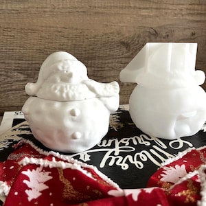 May include: Two white ceramic snowman-shaped containers. One has a lid and a scarf detail, the other has a hat. They are on a black surface with white text and a red and gold ribbon. The containers are likely for holiday decor.