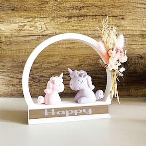 Può includere: Un arco in legno bianco con la parola "Happy" sulla base. Due piccole figurine di unicorno sono posizionate all'interno dell'arco. Un unicorno è rosa e l'altro è viola. Un mazzo di fiori secchi è posizionato sul lato destro dell'arco.