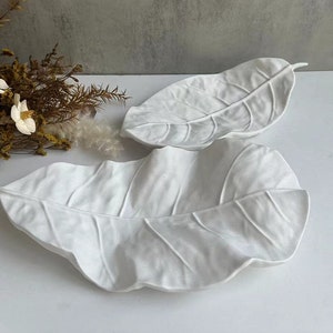 May include: Two white ceramic leaf-shaped serving dishes. The dishes are made to look like large leaves with detailed veins and a textured surface.