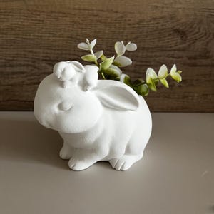 Könnte beinhalten: Ein weißer Keramik-Hasen-Pflanztopf mit einem kleineren Hasen auf dem Rücken, der grüne und weiße Kunstblätter hält. Der Hase hat lange Ohren.