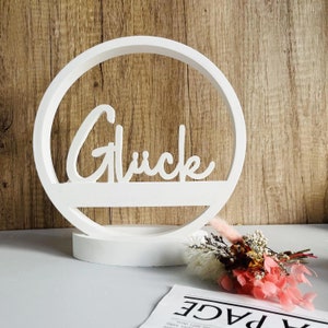 May include: White wooden circle with the word "Glück" in a cursive font. The circle is on a white base and there are dried flowers in front of it.