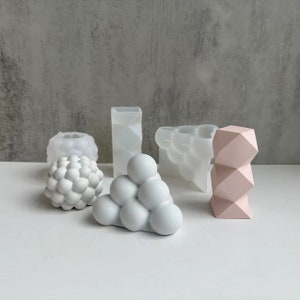 Puede incluir: Un conjunto de cinco macetas geométricas de hormigón blanco y rosa. Las macetas tienen diferentes formas, incluyendo una esfera, un cubo, un cilindro y un grupo de esferas.