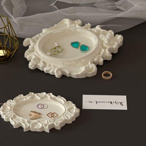 May include: Two white, ornate jewelry trays with various rings and necklaces. One tray holds a gold chain ring, two green heart-shaped earrings, and two rings. The other tray holds a necklace and two rings.