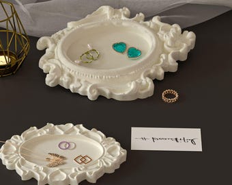 Puede incluir: Dos bandejas de joyería blancas y ornamentadas con varios anillos y collares. Una bandeja contiene un anillo de cadena dorada, dos pendientes verdes en forma de corazón y dos anillos. La otra bandeja contiene un collar y dos anillos.