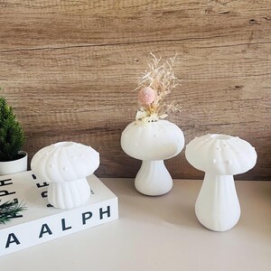 Puede incluir: Tres jarrones de cerámica blanca en forma de hongo con tapas texturizadas. El jarrón del centro tiene flores secas dentro.