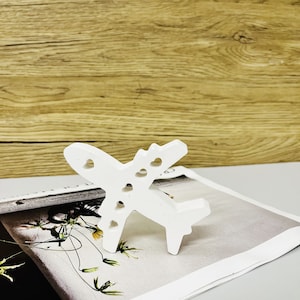 Puede incluir: Una figura de avión blanca con recortes en forma de corazón en las alas y el fuselaje. El avión decorativo se exhibe sobre una revista con un diseño floral, sobre un fondo de madera.