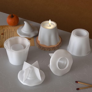 Pode incluir: Uma coleção de moldes e acessórios de velas brancas com tema de Halloween. Inclui uma vela em forma de fantasma acesa em uma base de madeira, um chapéu de bruxa e outros moldes. Uma pequena abóbora laranja também está presente.