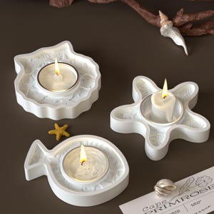 May include: Three white ceramic candle holders in the shapes of a seashell, starfish, and fish, each holding a lit tea light candle. A small starfish and a seashell are also present. The candle holders are on a brown surface.