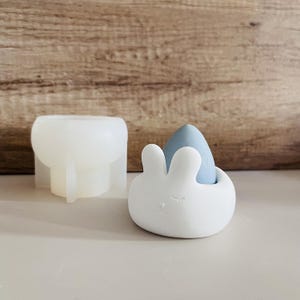 Op de afbeelding: Een witte keramische bunny-vormige houder met een lichtblauwe eivormige spons erin. De houder heeft een schattig gezicht met gesloten ogen en een kleine neus.