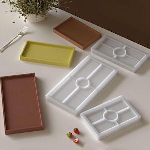 Op de afbeelding: Een assortiment rechthoekige trays in verschillende kleuren, waaronder bruin, geel en transparant. De transparante trays hebben een verdeeld ontwerp. Verse aardbeien zijn bij de trays geplaatst. Een vork en een kleine plant in een witte pot zijn ook zichtbaar.