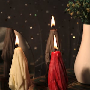 May include: Three candles shaped like draped fabric, one is white, one is red, and one is grey. The candles are lit and the flames are visible.