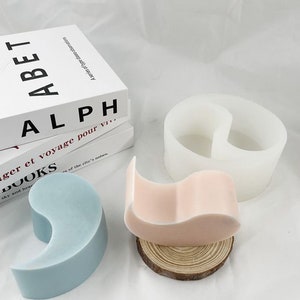 May include: Two silicone molds for making resin crafts. One mold is a yin yang shape and the other is a heart shape. The molds are white and are sitting on a wooden surface.