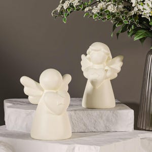 May include: Two off-white angel figurines, each holding a heart with a star. The angels have wings and are standing on a white, textured surface. A vase with white flowers is in the background.