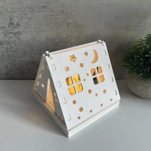 Puede incluir: Luz nocturna de madera blanca con forma de casa con un diseño recortado de estrellas, una luna creciente y ventanas. La luz está encendida y brillando.