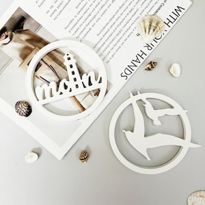 Puede incluir: Dos piezas de decoración de pared de madera blanca con temática náutica. Una pieza presenta un faro y la palabra "mom" en una fuente estilizada. La otra pieza presenta dos gaviotas estilizadas en vuelo.