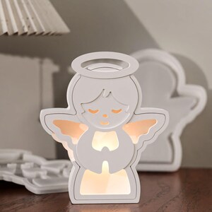 Puede incluir: Una luz de noche blanca en forma de ángel con un halo, manos juntas en oración y alas brillantes. La luz tiene un brillo suave y cálido. Un objeto similar en forma de ángel está en el fondo.