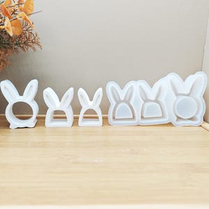 May include: A collection of white bunny-shaped molds and forms, perfect for crafting. The set includes various sizes, from a ring-shaped bunny to a mold with multiple bunny shapes. The items are arranged on a light brown wooden surface.