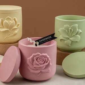 May include: Three decorative candle holders in pastel colors. The yellow holder has a lotus flower design, the green holder has a lily design, and the pink holder has a rose design. The pink holder has a lid and a tag that says "HANDMADE Especially for You."