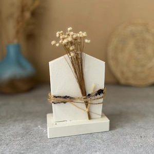 Puede incluir: Un jarrón de cerámica blanca con forma de casa con un arreglo de flores secas. El jarrón está atado con cuerda y se asienta sobre una base blanca.