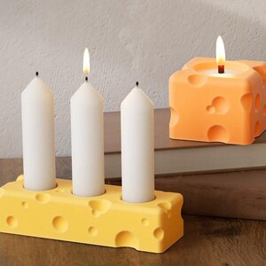 May include: Four white candles in a yellow cheese-shaped candle holder. A single orange cheese-shaped candle sits on a stack of books.
